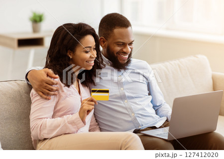 Online Shopping, Ecommerce Concept. Happy young african american couple using laptop and buying clothes or products on internet, paying with debit credit card, sitting on the sofa at home 127412020