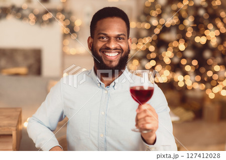 Celebration Concept. Portrait of smiling handsome young bearded african american man holding glass of red wine, toasting and looking to camera, blurred background, selective focus 127412028