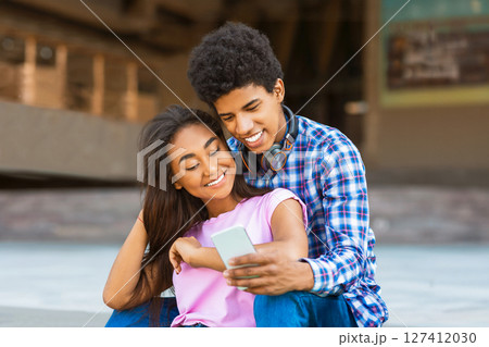 A teen black couple sits together on a concrete surface in an urban environment. Guy is wearing a blue and white plaid shirt and jeans, while the girl wears a pink T-shirt. They both appear happy A teen black couple sits together on a concrete surface in an urban environment. Guy is wearing a blue and white plaid shirt and jeans, while the girl wears a pink T-shirt. They both appear happy 127412030