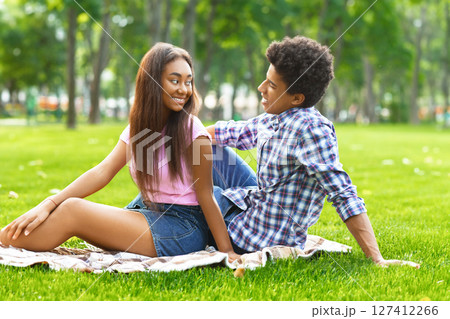 A teen black couple sits on a blanket in a park on a sunny day. The girl is wearing a pink shirt and a denim skirt and is looking at guy. Guy is wearing plaid shirt and jeans and is looking at girl 127412266