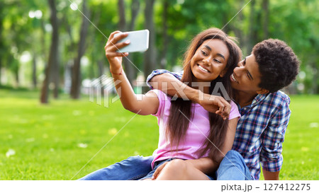 A teen black couple sits on a grassy lawn in a park on a sunny day. The girl is taking a selfie with her phone while guy smiles at her and puts his arm around her waist. 127412275