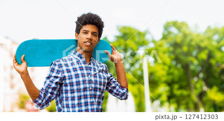 A teen black guy with dark curly hair and a plaid shirt is smiling while carrying a blue skateboard on his shoulder. The background shows a blurred view of trees and buildings, copy space 127412303