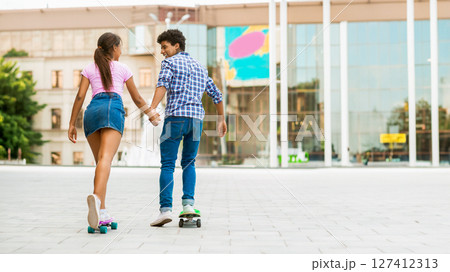 A teen black couple, a girl in a pink shirt and blue skirt, and a boy in a plaid shirt and blue jeans, skateboard side by side in a modern city setting. They are holding hands, copy space A teen black couple, a girl in a pink shirt and blue skirt, and a boy in a plaid shirt and blue jeans, skateboard side by side in a modern city setting. They are holding hands, copy space 127412313