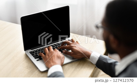 Concentrated black businessman typing on laptop with blank screen, working in office. Over shoulder shot Concentrated black businessman typing on laptop with blank screen, working in office. Over shoulder shot 127412370