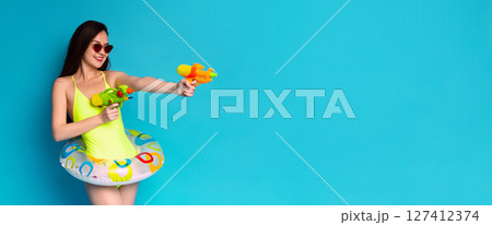 A woman in a bright yellow swimsuit and sunglasses holds two water guns, one in each hand. She has a playful expression on her face and is looking towards the right side of the frame A woman in a bright yellow swimsuit and sunglasses holds two water guns, one in each hand. She has a playful expression on her face and is looking towards the right side of the frame 127412374