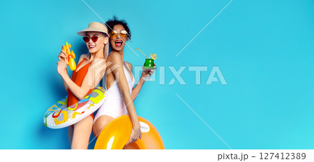 This image features two women in swimsuits posing against a blue background. Both women are smiling and look ready for a fun day at the pool, copy space 127412389