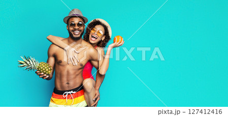 African American young couple is seen smiling and having fun in front of a bright turquoise background. The man wearing swim trunks and a hat, while the woman is in a swimsuit and holding an orange. 127412416
