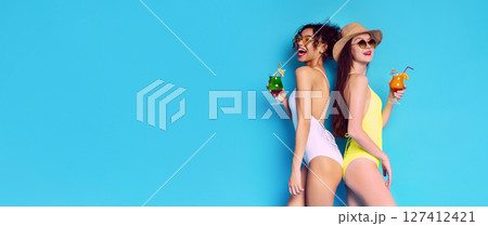 This image shows two women in swimsuits, standing back-to-back and smiling. They are both holding cocktails and appear to be enjoying a summer day, copy space 127412421