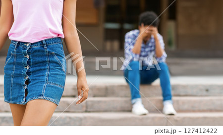 A girl in a pink shirt and denim skirt walks away from a guy sitting on concrete steps. Guy is wearing jeans and a plaid shirt. The girl is looking straight ahead while guy is looking down A girl in a pink shirt and denim skirt walks away from a guy sitting on concrete steps. Guy is wearing jeans and a plaid shirt. The girl is looking straight ahead while guy is looking down 127412422