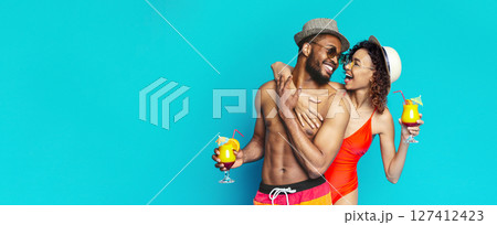 African American couple in swimwear, a man wearing a hat and a woman wearing a red swimsuit, are laughing and holding cocktails in front of a bright blue background, copy space 127412423