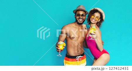 African American young couple, dressed in summer attire, smiles brightly while enjoying cocktails. They are standing in front of a bright blue background, possibly a studio backdrop. African American young couple, dressed in summer attire, smiles brightly while enjoying cocktails. They are standing in front of a bright blue background, possibly a studio backdrop. 127412431