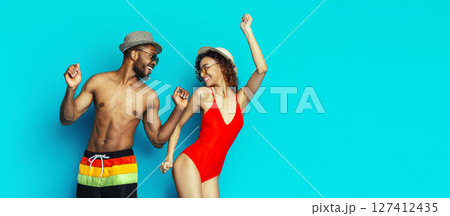 Black man and woman are joyfully dancing in front of a bright blue background. The man is wearing swim trunks and a hat, while the woman is wearing a red swimsuit and a hat. Black man and woman are joyfully dancing in front of a bright blue background. The man is wearing swim trunks and a hat, while the woman is wearing a red swimsuit and a hat. 127412435