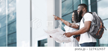 African American couple stand outside a building with modern architecture. The woman is pointing to the right, while the man is holding a map and smiling, copy space 127412437