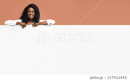A young black woman with long, dark curly hair leaning on large white sign in front of a peach-colored background. She is smiling and looking directly at the camera 127412443