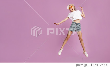 A young woman is captured mid-air, jumping with excitement against a pink backdrop. She is wearing a white t-shirt, denim shorts, and sunglasses, and is smiling broadly with her arms outstretched. A young woman is captured mid-air, jumping with excitement against a pink backdrop. She is wearing a white t-shirt, denim shorts, and sunglasses, and is smiling broadly with her arms outstretched. 127412453