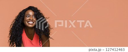 The image shows a young black woman with curly black hair smiling and looking upwards. She is wearing a red tank top and has a natural, healthy look. The background is a solid peach color. The image shows a young black woman with curly black hair smiling and looking upwards. She is wearing a red tank top and has a natural, healthy look. The background is a solid peach color. 127412463