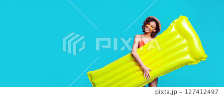 A young African American woman with curly hair and sunglasses is smiling and holding a large yellow inflatable raft against a blue background, copy space A young African American woman with curly hair and sunglasses is smiling and holding a large yellow inflatable raft against a blue background, copy space 127412497