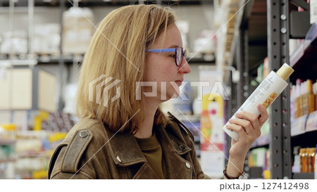 Young woman wearing glasses choosing cosmetics, reading label on bottle of shampoo or hair conditioner in supermarket aisle 127412498