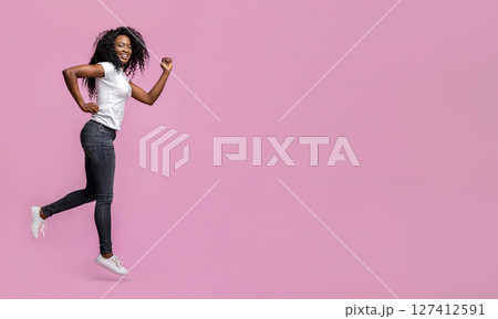 A young Black woman with curly hair is joyfully jumping in the air in front of a pink background. She is wearing a white t-shirt and dark jeans, and she has a bright smile on her face. A young Black woman with curly hair is joyfully jumping in the air in front of a pink background. She is wearing a white t-shirt and dark jeans, and she has a bright smile on her face. 127412591