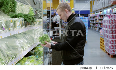 Man carefully selects fresh vegetables in a brightly lit supermarket aisle, surrounded by green produce, showcasing healthy eating choices. Man choosing greens in supermarket, shelf with greens Man carefully selects fresh vegetables in a brightly lit supermarket aisle, surrounded by green produce, showcasing healthy eating choices. Man choosing greens in supermarket, shelf with greens 127412625