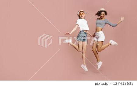 This image shows two young women jumping in the air in front of a pink background. They are both smiling and appear to be having fun, copy space This image shows two young women jumping in the air in front of a pink background. They are both smiling and appear to be having fun, copy space 127412635