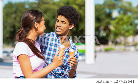 A teen black couple stand close together and smile as they each hold a waffle cone ice cream. They are standing outside on a sunny day, copy space 127412641