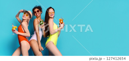 Three women in swimsuits are standing in front of a blue background. They are all smiling and holding cocktails. The women appear to be enjoying themselves on a summer day. Three women in swimsuits are standing in front of a blue background. They are all smiling and holding cocktails. The women appear to be enjoying themselves on a summer day. 127412654