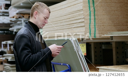 A man in a hardware store, choosing lumber, shelves with lumber. Customer uses a smartphone to compare prices, check availability, and find information on construction materials at a hardware store A man in a hardware store, choosing lumber, shelves with lumber. Customer uses a smartphone to compare prices, check availability, and find information on construction materials at a hardware store 127412655