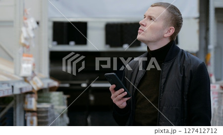 Contractor browsing building materials on a smartphone in a lumber yard or home improvement store. A man in a hardware store, choosing flooring, shelves with ceramic tiles. 127412712