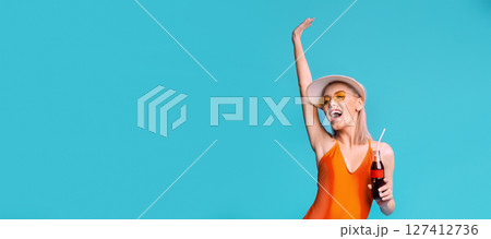 A woman in an orange swimsuit smiles and holds a soda while wearing a white sun hat and yellow sunglasses. She stands against a solid blue background with her arm raised, copy space A woman in an orange swimsuit smiles and holds a soda while wearing a white sun hat and yellow sunglasses. She stands against a solid blue background with her arm raised, copy space 127412736