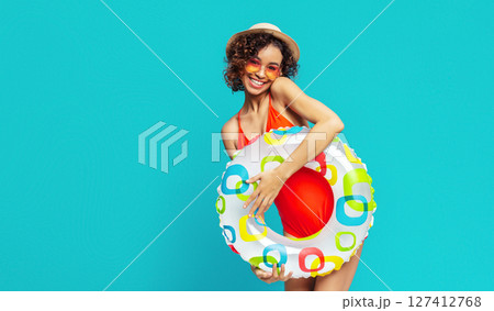 Young black woman with curly brown hair, wearing a red swimsuit, sunglasses, and a straw hat, holding a colorful inflatable ring against a bright blue background. 127412768