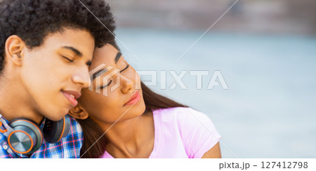 A teen African American couple rests their heads together, eyes closed, while outdoors. The background is a blurred image of a light blue surface, panorama with copy space 127412798