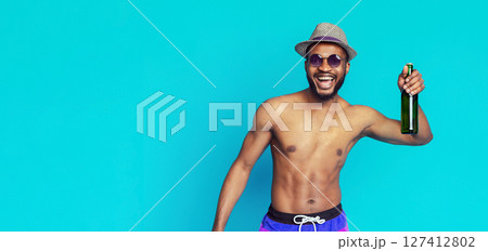 African American smiling man wearing a straw hat and sunglasses, with his torso bare and wearing swim trunks, holding a green glass beer bottle in front of a blue background. 127412802