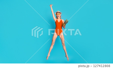 Pretty young blonde woman in red swimsuit, sunglasses and summer hat holding pineapple and gesturing while jumping in the air over blue studio background Pretty young blonde woman in red swimsuit, sunglasses and summer hat holding pineapple and gesturing while jumping in the air over blue studio background 127412808