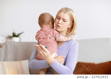 Concerned young mother looking at electronic thermometer, her little kid got sick, home interior. Worried blonde lady with toddler in her arms looking at thermometer. Baby health, illness concept 127412815