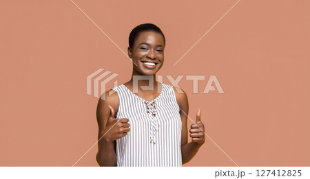 A young Black woman with short hair is smiling and giving a thumbs up with both hands. She is wearing a white and black striped sleeveless top and standing in front of a peach-colored background. A young Black woman with short hair is smiling and giving a thumbs up with both hands. She is wearing a white and black striped sleeveless top and standing in front of a peach-colored background. 127412825