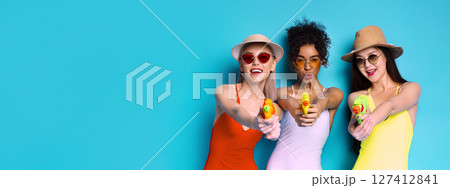 Three women are standing in front of a blue background. They are all wearing sunglasses and hats, and they are holding water guns, copy space Three women are standing in front of a blue background. They are all wearing sunglasses and hats, and they are holding water guns, copy space 127412841