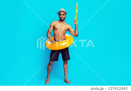 Black man wearing a hat, sunglasses, and swim trunks stands in front of a blue background. He is holding a yellow water gun in his right hand and an inflatable ring around his waist. Black man wearing a hat, sunglasses, and swim trunks stands in front of a blue background. He is holding a yellow water gun in his right hand and an inflatable ring around his waist. 127412842