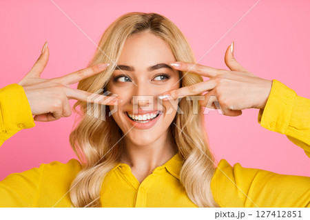 A blonde woman with long, wavy hair smiles and poses in front of a pink background. She is wearing a yellow shirt and has her fingers forming peace signs in front of her eyes. A blonde woman with long, wavy hair smiles and poses in front of a pink background. She is wearing a yellow shirt and has her fingers forming peace signs in front of her eyes. 127412851