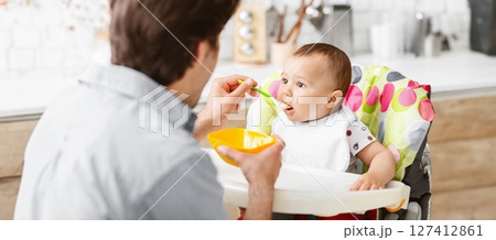 A father feeds his baby son sitting in a high chair with a spoon. The baby is wearing a white bib and has a bowl of food in front of him. The father is looking at the baby with a loving expression 127412861