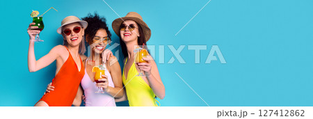 Three friends, wearing swimsuits, hats, and sunglasses, smile brightly as they hold cocktails in front of a blue background. The woman on the left holds a green cocktail with a straw, copy space 127412862