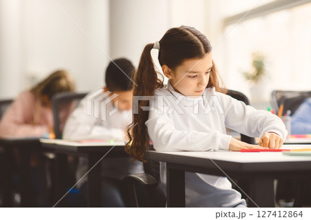 Portrait of small girl with ponytails sitting at table in classroom at primary school or kindergarten, writing or drawing in notebook. Reopening and return back to school after coronavirus quarantine Portrait of small girl with ponytails sitting at table in classroom at primary school or kindergarten, writing or drawing in notebook. Reopening and return back to school after coronavirus quarantine 127412864