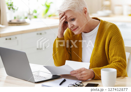 Upset senior lady holding her head while checking bills, having financial troubles during quarantine. Sad elderly woman sitting in kitchen in front of laptop and holding documents, copy space 127412877