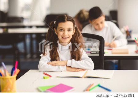 First Day At School. Portrait of smiling diligent small girl with ponytails sitting at table in classroom at elementary school with group of classmates in background and posing, looking at camera 127412881