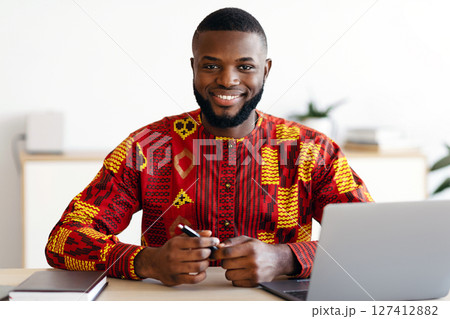 Handsome African Freelancer Guy In Traditional Costume Sitting At Desk At Home Office, Working On Laptop Computer And Smiling At Camera, Black Man Enjoying Remote Work And Freelance Career, Free Space 127412882
