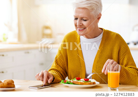 Happy old lady modern grandmother using smartphone while having lunch, eating sandwich and drinking juice, sitting alone at kitchen, empty space. Senior woman reading blog on mobile phone 127412883