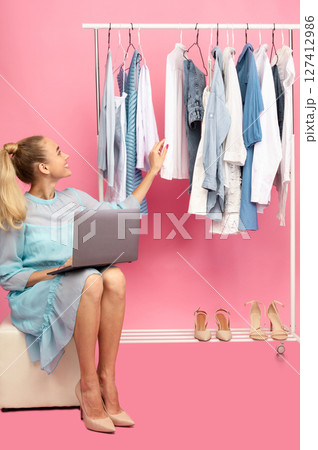 Fashion Designer. Vertical portrait of happy stylish lady sitting in closet on pouffe with computer on lap near rack with clothes and shoes, choosing outfit, isolated on pastel pink studio background Fashion Designer. Vertical portrait of happy stylish lady sitting in closet on pouffe with computer on lap near rack with clothes and shoes, choosing outfit, isolated on pastel pink studio background 127412986