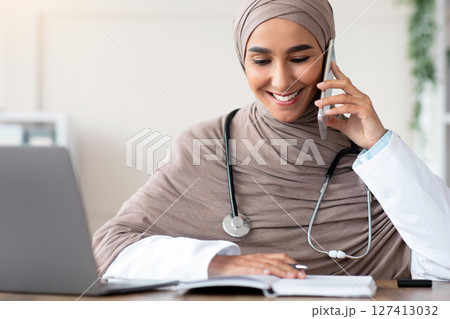 Smiling young lady in hijab doctor talking on mobile phone and checking her notebook, making appointment, sitting at workdesk in front of laptop at clinic, copy space. Remote healthcare, telehealth 127413032