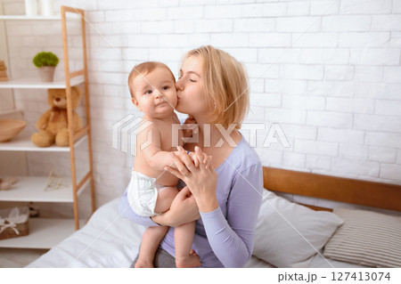 Young blonde mother kissing her adorable little kid son or daughter, bedroom interior, free space. Loving mom and cute baby boy or girl in diaper, mummy waking up her sweet baby 127413074
