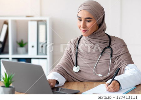 Young woman doctor having online appointment, sitting in office, using laptop and taking notes, copy space. Attractive muslim lady physician working with computer, clinic interior, remote healthcare 127413111